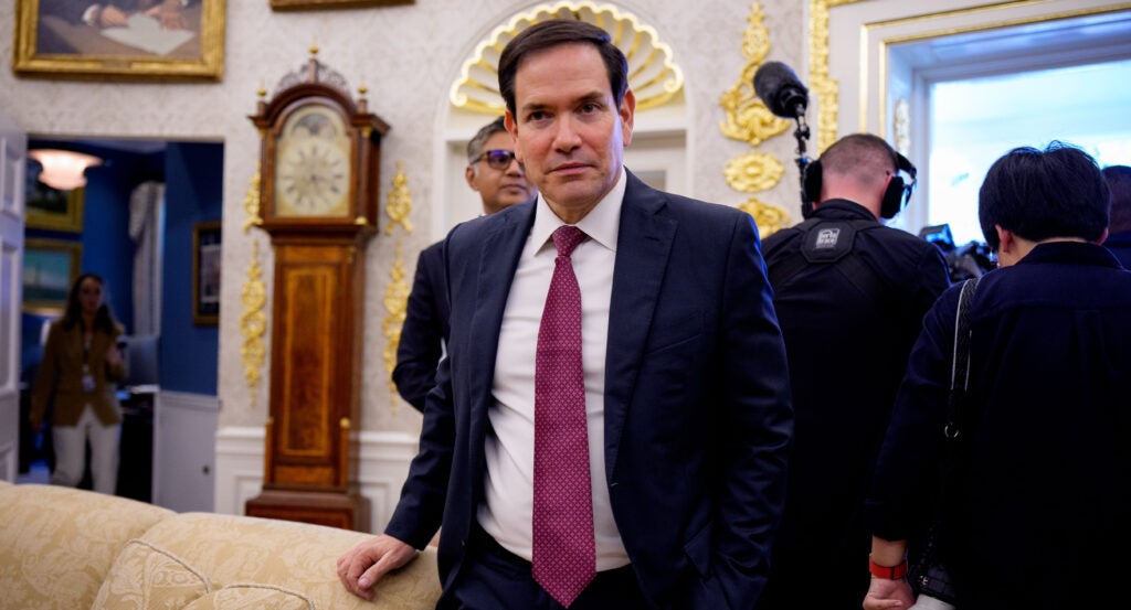 Secretary of State Marco Rubio is standing near a sofa, looking directly at the camera.