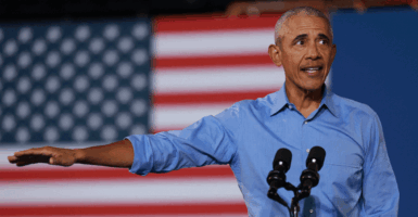 Former President Barack Obama at rally in in Clarkston, Georgia for 2024 Democratic presidential nominee, Kamala Harris. Obama is on stage in front of an American Flag wearing a blue shirt with his right arm raised pointing to his right.