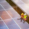 Aerial view of two solar energy technicians examining large solar panels in a field.