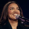 Alexandria Ocasio-Cortez smiles as she stands before a microphone.