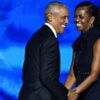 Michelle Obama and Barack Obama hold each other's hands as they stand together on a stage while smiling.