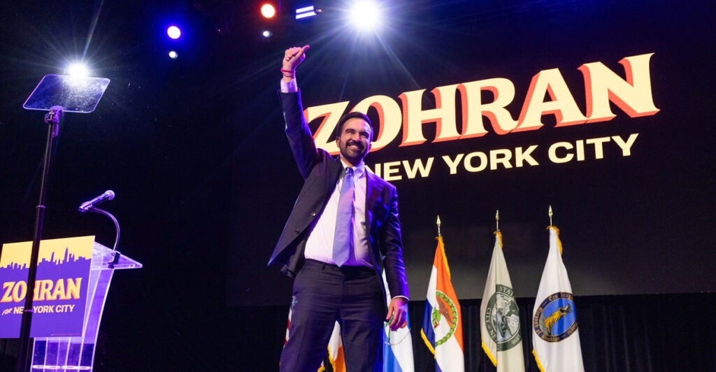 Zohran Mamdani gives a thumbs up sign with his right hand as he walks across a stage with a smile on his face.