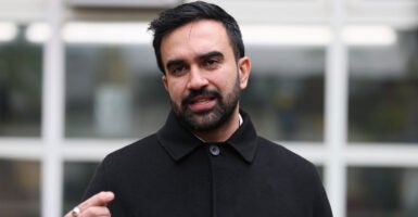 Zohran Mamdani stands as he talks to reporters with his head tilted to his right while motioning with his right hand.