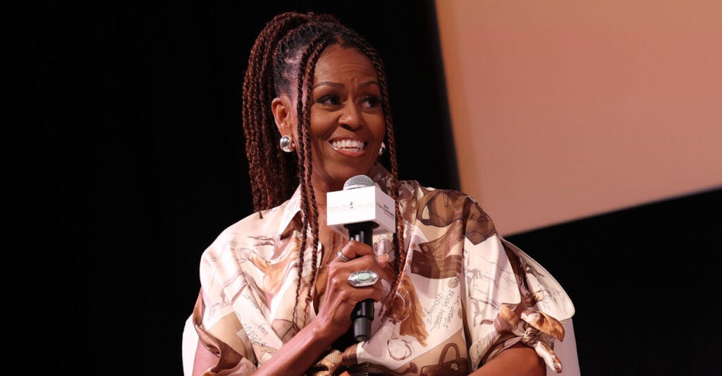 Michelle Obama sits in a chair and holds a microphone in her right hand as she turns to the left.