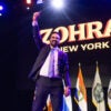 Zohran Mamdani raises a fist on a stage backed with a sign reading "ZOHRAN New York City."