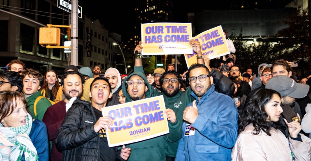 A group of Zohran Mamdani supporters, some carrying "Our Time Has Come" signs, cheer his victory.