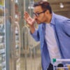 Man in blue shirt, white T-shirt, and glasses appears to be yelling at items in grocery store fridge.