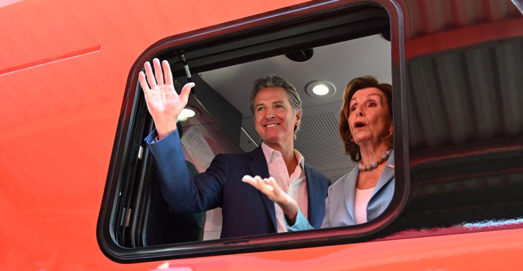 Gavin Newsom and Nancy Pelosi wave from a red electric train car.