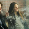 A young male and female sit along a brick wall vaping.