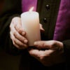 Hands of a priest holding a lit white candle.