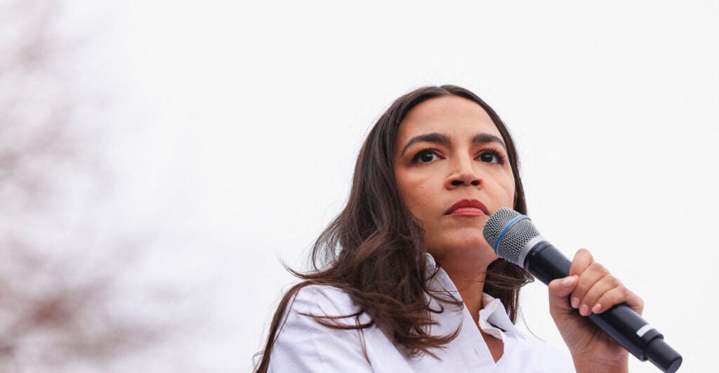 Alexandria Ocasio-Cortez in a white blouse behind a microphone, with a pale, outdoor background.