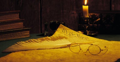 Spectacles and quill pen lay atop a copy of the U.S. Constitution next to a lit candle.