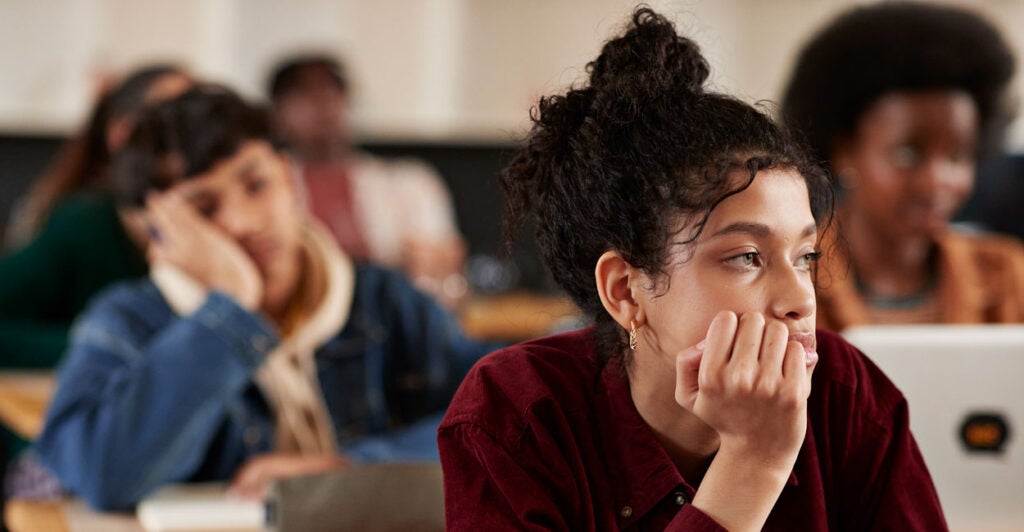 Bored teenage female looks out classroom window while male classmate behind her is fast asleep.