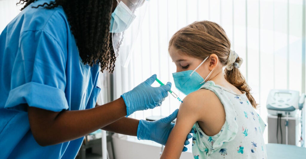 A young girl in a mask receives a COVID-19 vaccine shot in her left arm from a masked nurse.