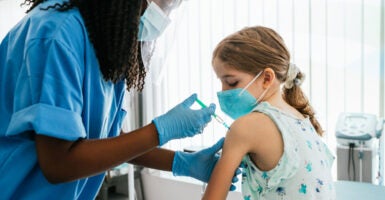A young girl in a mask receives a COVID-19 vaccine shot in her left arm from a masked nurse.