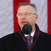 Veterans Affairs Secretary Doug Collins in a winter overcoat speaks in front of a large American flag.
