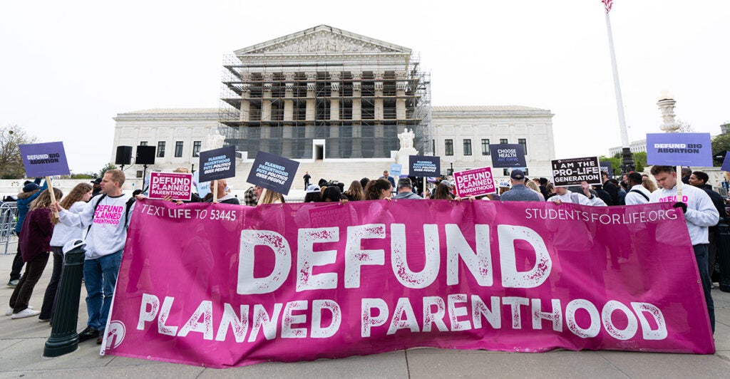 Pro-life protesters in front of the Supreme Court with pink sign reading "Defund Planned Parenthood"