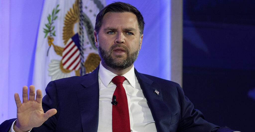 JD Vance seated, in a blue suit with red tie, with his right hand up.