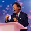 Tucker Carlson with fists clenched speaks at pink podium.