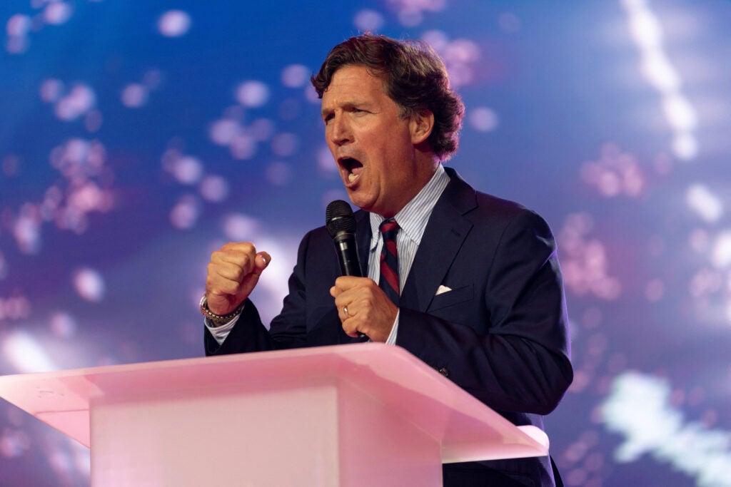 Tucker Carlson with fists clenched speaks at pink podium.