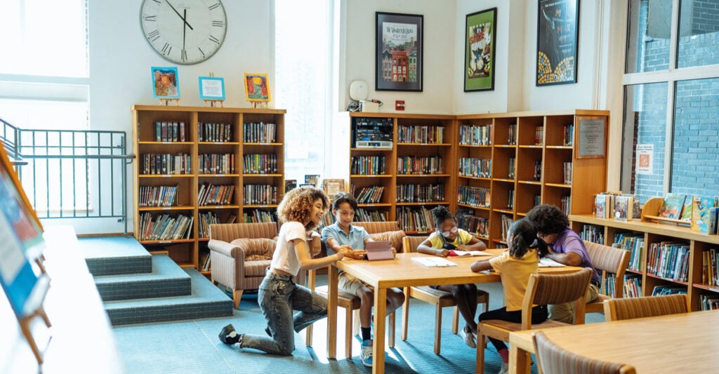 A woman kneels at a table where a young child is reading a book in a library.