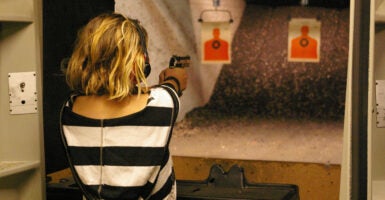 Blonde woman in black and white shirt fires handgun at an indoor shooting range.