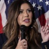 Close-up of Kristi Noem holding microphone and pointing while standing in front of American flag.