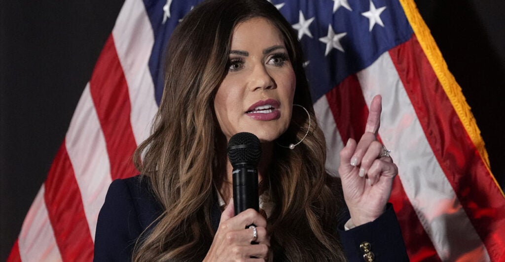 Close-up of Kristi Noem holding microphone and pointing while standing in front of American flag.