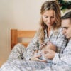 Parents in grey and white pajamas sit on their bed, holding a newborn. A Christmas tree is on the left side of the shot.