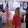 Little children stand in their classroom in front of a boy holding an American flag.