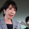 Japanese Prime Minister Sanae Takaichi, in a grey jacket and black shirt, speaks into bank of microphones.