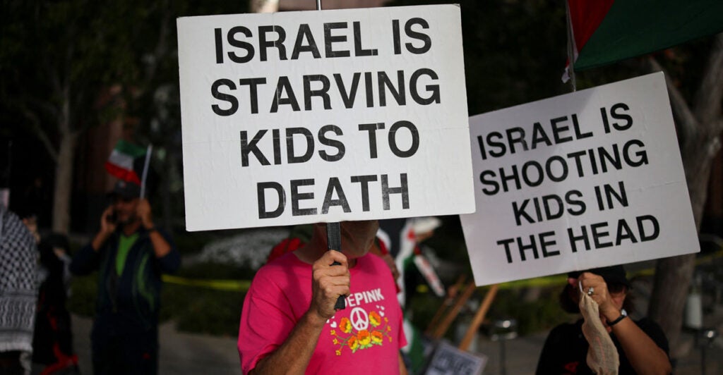Anti-Israel protesters hold signs reading "Israel is Starving Kids to Death" and "Israel Shoots Kids in the Head" during rally.