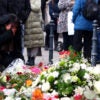 A woman grieves at a flower-strewn memorial as people stand around.