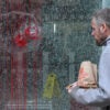 A man in grey jacket walks past a shattered window that's covered in fake blood.