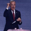 Canadian Prime Minister Mark Carney with his right fist raised and left hand holding tag that reads "Canada" stands at a pink podium.