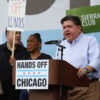 JB Pritzker in a white shirt speaks at a podium that has a sign "Hands Off Chicago."