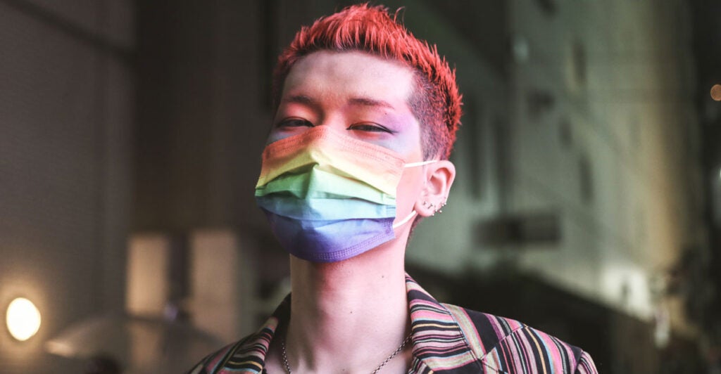 A thin young man with dyed red hair, makeup and a rainbow mask.
