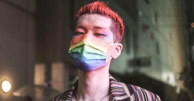 A thin young man with dyed red hair, makeup and a rainbow mask.