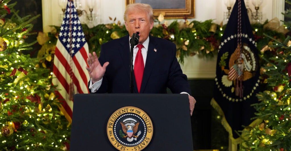 President Trump behind a White House podium, with Christmas garland and an American flag behind him, gestures with his right hand.