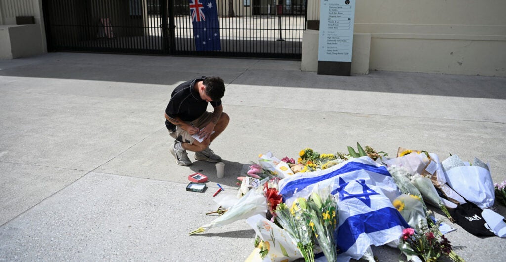 Man kneels while writing at edge of memorial for Bondi Beach Hanukkah massacre victims.