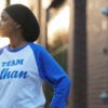 Ihan Omar in a blue-sleeved jersey reading "Team Ilhan" stands on a street.