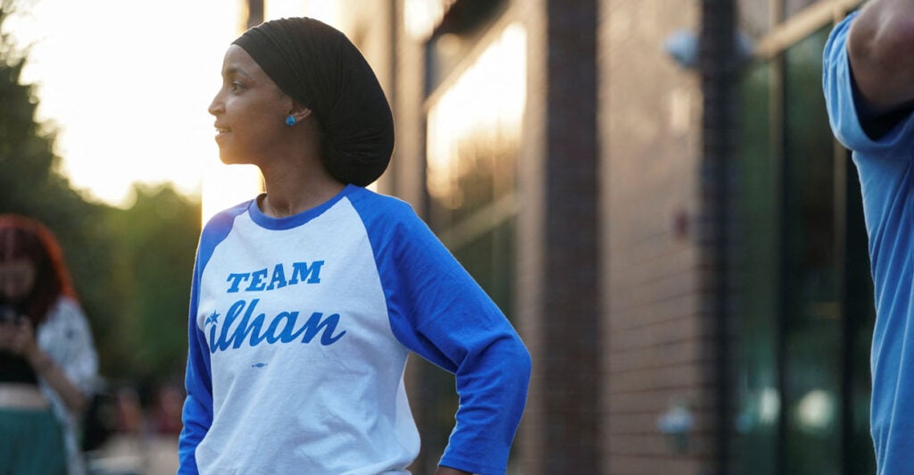 Ihan Omar in a blue-sleeved jersey reading "Team Ilhan" stands on a street.