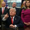President Trump points to a reporter while seated at his Oval Office desk surrounded by officials.