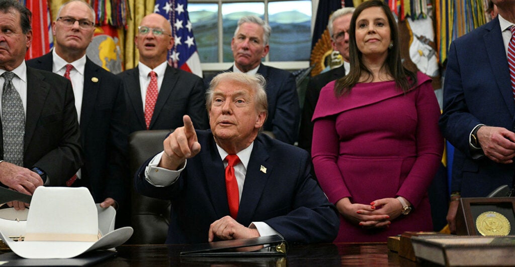 President Trump points to a reporter while seated at his Oval Office desk surrounded by officials.