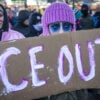 A protester with purple knit hat and blue makeup around eyes holds sign reading "ICE OUT"