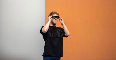 Mark Zuckerberg, in a black t-shirt, puts on a pair of dark glasses against a half-white, half-orange background.