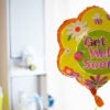 A yellow balloon with red lettering spelling "Get Well Soon" floats in a hotel room.