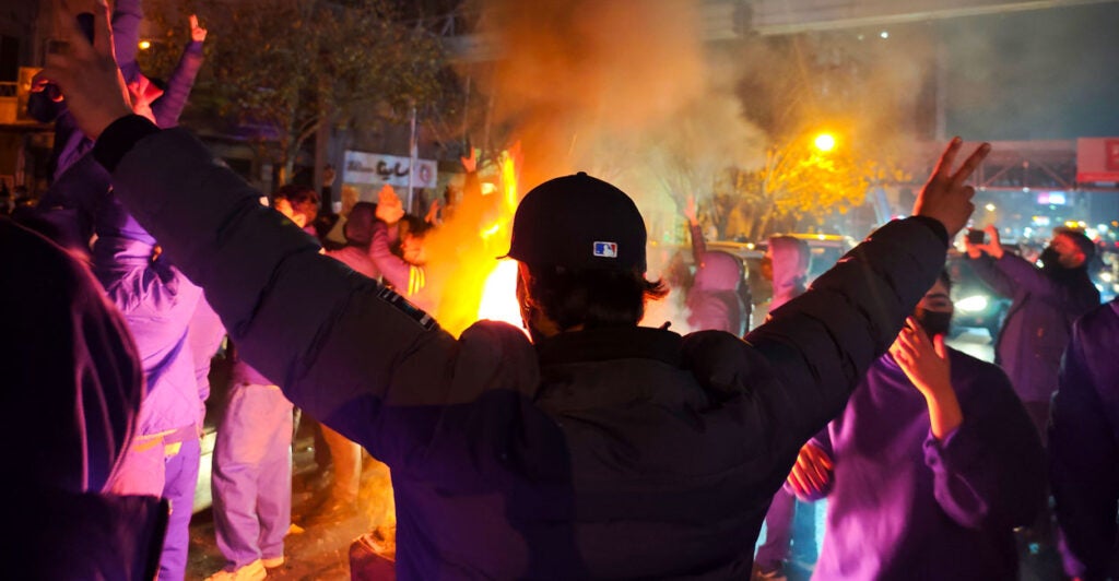 Iranian protester with back to the camera, arms outstretched, giving peace signs, amid nighttime protest. Flames in front of him.