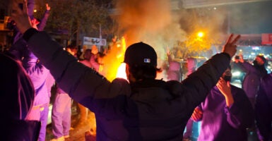 Iranian protester with back to the camera, arms outstretched, giving peace signs, amid nighttime protest. Flames in front of him.