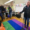 Gavin Newsom addresses the press while in an elementary school classroom, standing on a rainbow -colored rug.